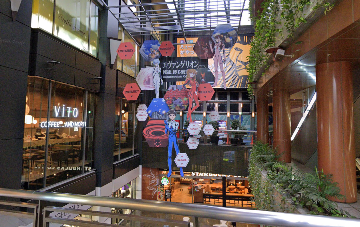A photo taken on a bridge between two parts of Canal City towards a display of Evangelion characters. Shown are Shinji, Asuka, Rei, and a fourth character I don't know the identity of because I've never watched Evangelion. Hexagonal signs saying HAKATA are displayed in the style of Evangelion's EMERGENCY warnings.