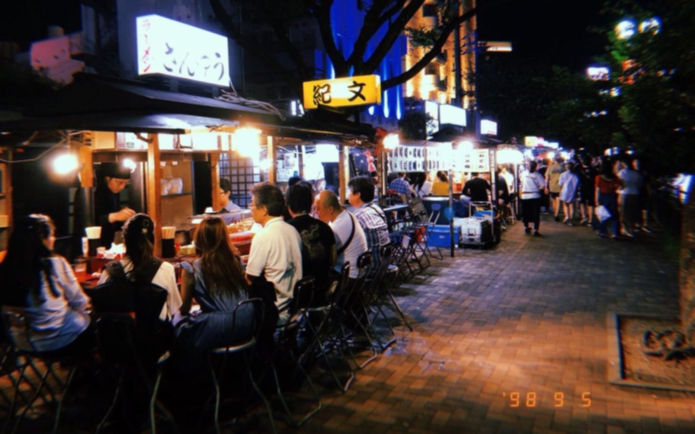 A photo of Yatai in Fukuoka. It's late at night, and the signs of many food stands illuminate the sidewalk. There's many patrons at each stand.