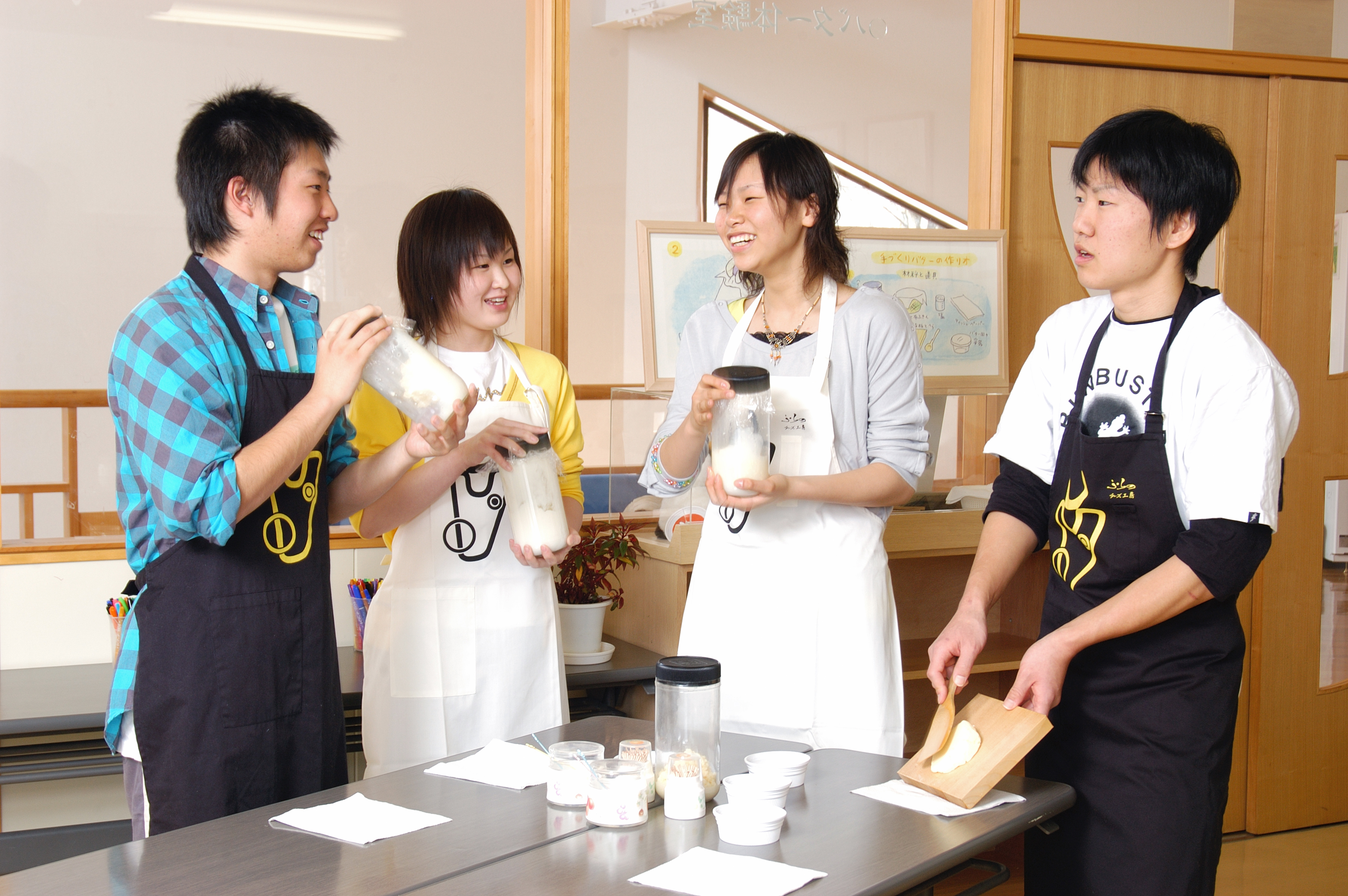 Four people taking part in a butter-making workshop. Three are shaking jars with dairy inside to create butter, and the fourth has a lump of butter on a wooden block in one hand and a butter paddle in the other.