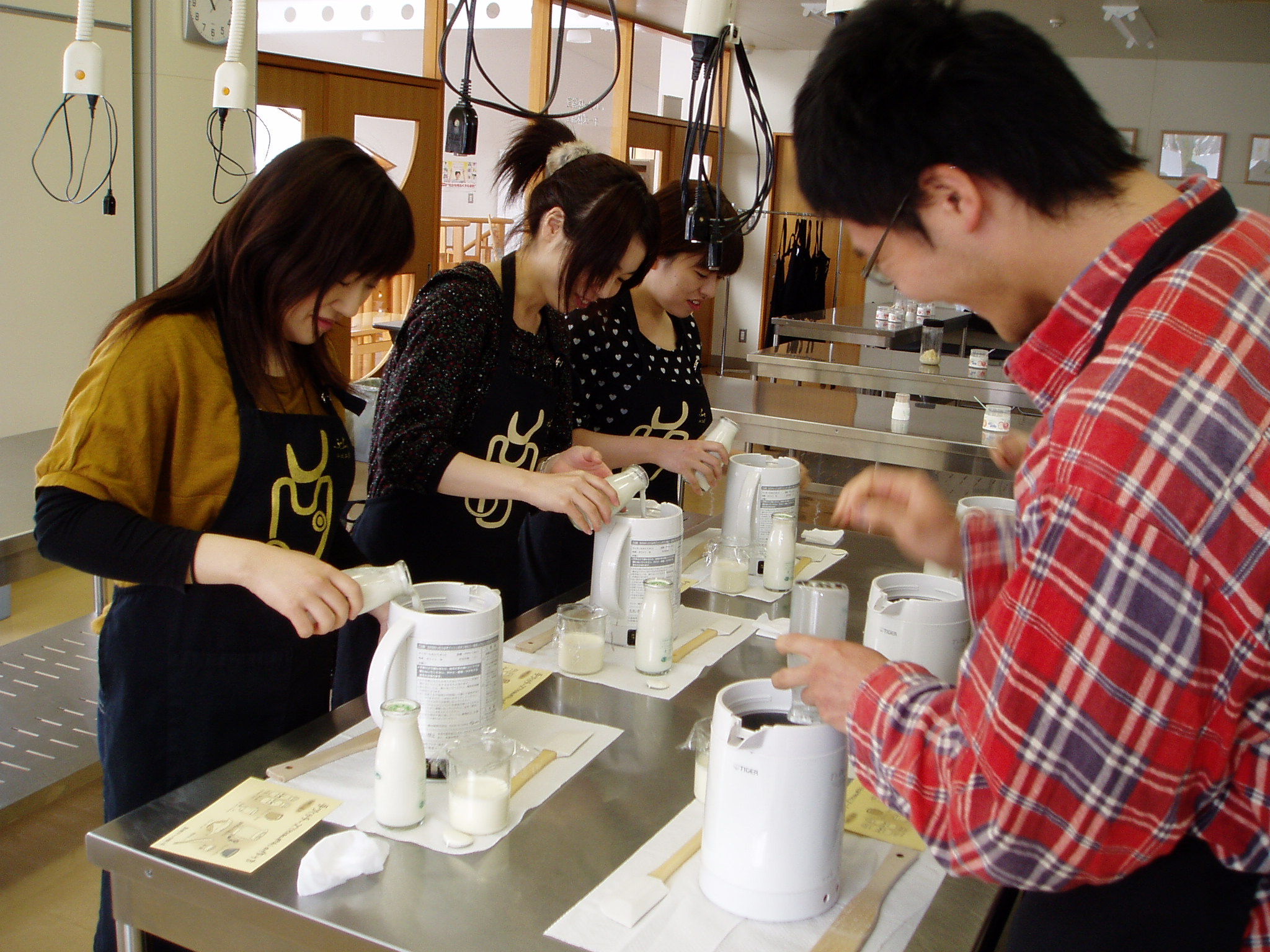 Five or six (unclear) people taking part in a cheese-making workshop. At the present moment, they're pouring some milk-like product into some machine.