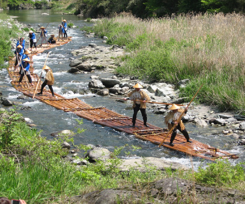 A chain of 16 rafts, with a Japanese person standing on 10 of them. Almost everyone is wearing a kasa. They are paddling down a river using some sort of long stick.