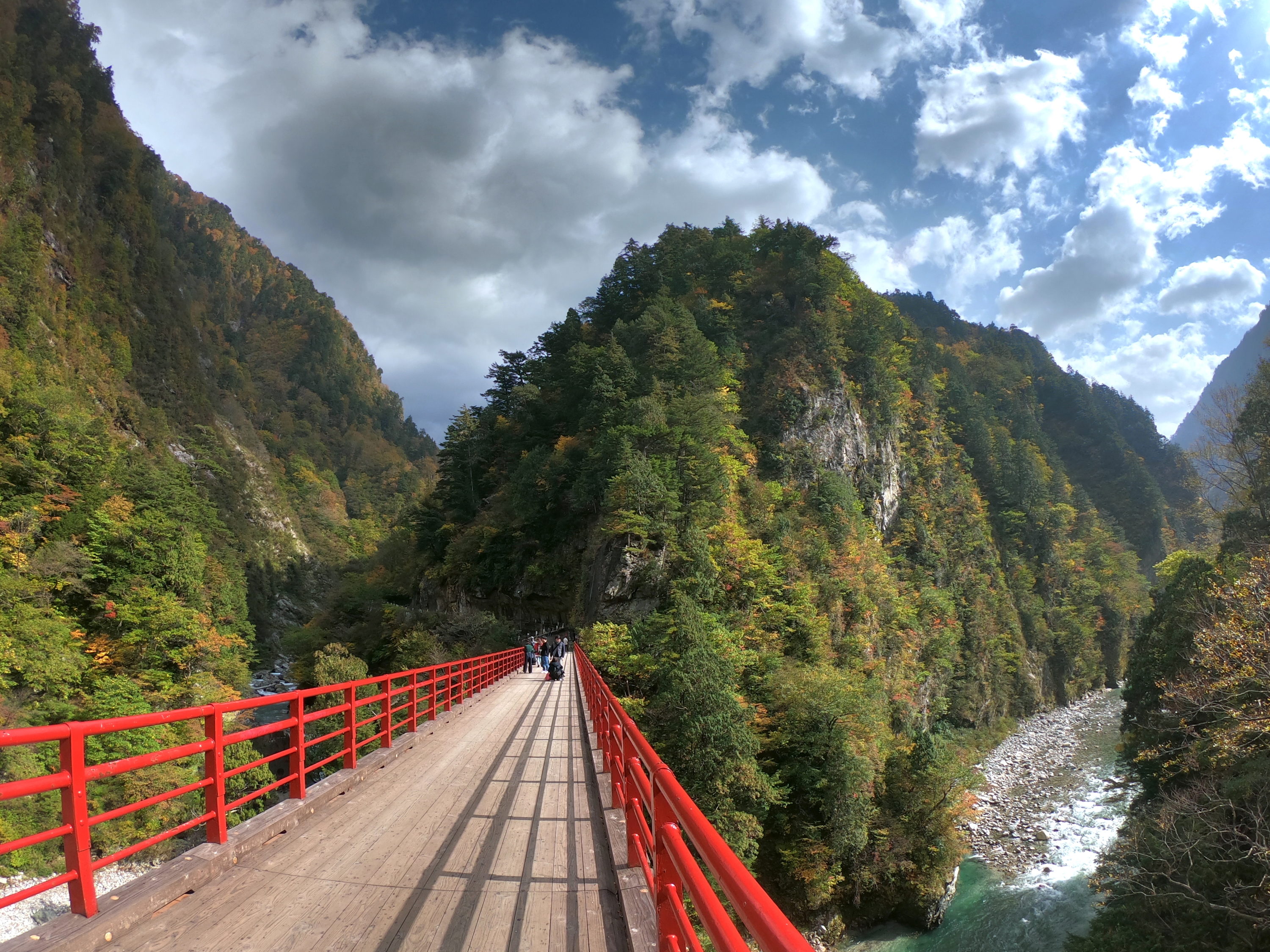 A photo taken on one end of a red bridge crossing a gorge with a flowing river underneath. On both sides of the gorge there are beautiful green trees.