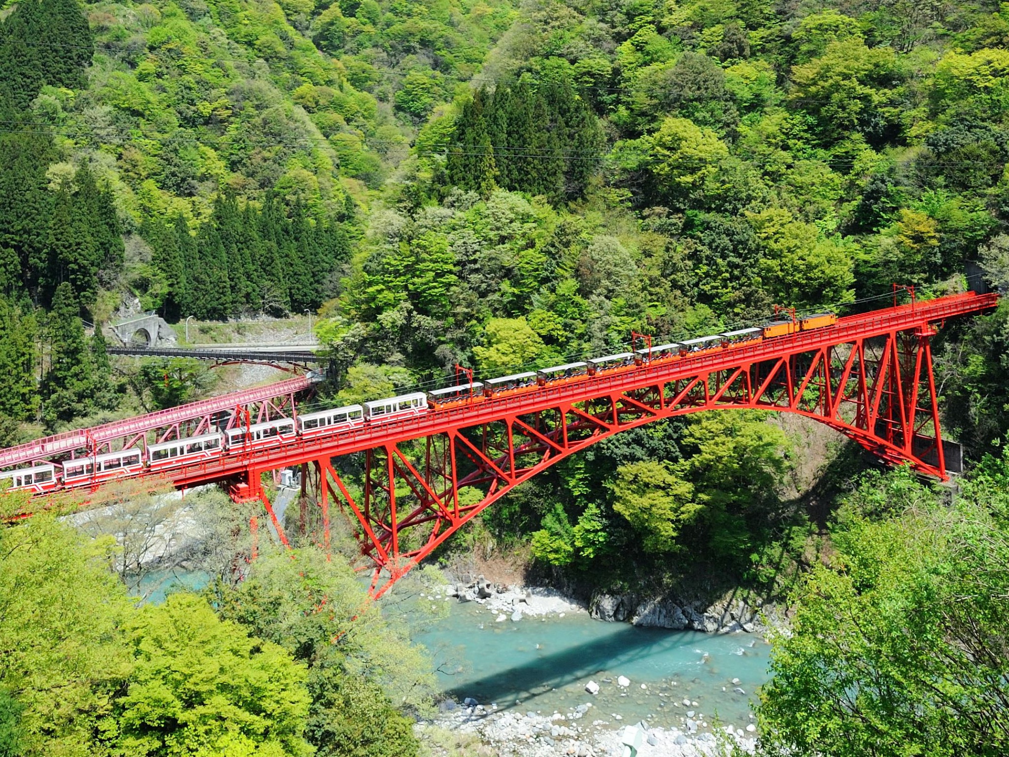 A red bridge is running over a blue river. Steep hills, not a single spot without tall, lush trees, line the area. A tourism trolley runs along the tracks on the bridge.