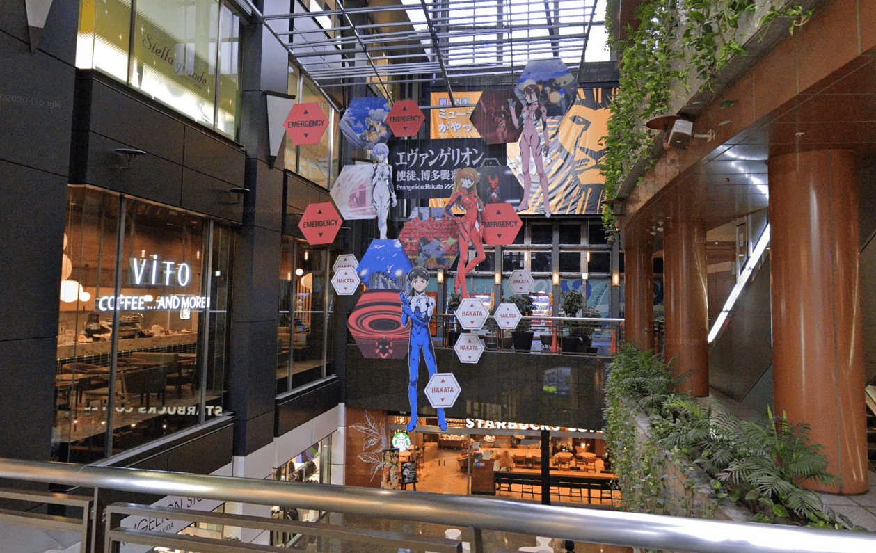 A photo taken on a bridge between two parts of Canal City towards a display of Evangelion characters. Shown are Shinji, Asuka, Rei, and a fourth character I don't know the identity of because I've never watched Evangelion. Hexagonal signs saying HAKATA are displayed in the style of Evangelion's EMERGENCY warnings.